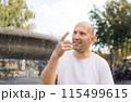 Bald man in white t-shirt smiling, pointing near fountain, outdoor park setting 115499615