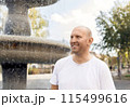 Bald man in white t-shirt smiling, pointing near fountain, outdoor park setting 115499616