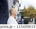 Bald man in white t-shirt leaning against wall, serious expression, urban background 115499619