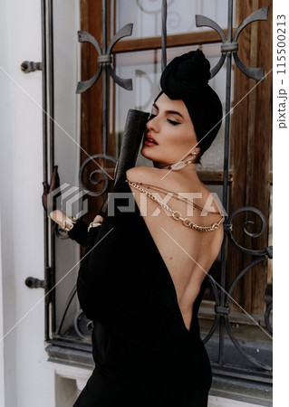 Stylish woman in the city. Fashion photo of a beautiful model in an elegant black dress posing against the backdrop of a building on a city street 115500213