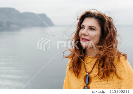 Portrait windswept hair happy woman against a backdrop of mountains and sea. Daylight illuminates the tranquil outdoor setting 115500231