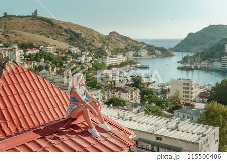 Woman sits on rooftop, enjoys town view and sea mountains. Peaceful rooftop relaxation. Below her, there is a town with several boats visible in the water. Rooftop vantage point. 115500406
