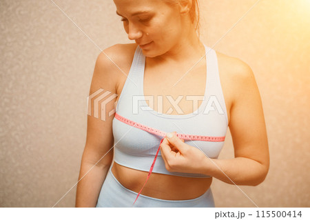 Cropped view of slim woman measuring breasts with tape measure at home, close up. A European woman checks the result of a weight loss diet or liposuction indoors. Healthy lifestyle. Cropped view of slim woman measuring breasts with tape measure at home, close up. A European woman checks the result of a weight loss diet or liposuction indoors. Healthy lifestyle. 115500414