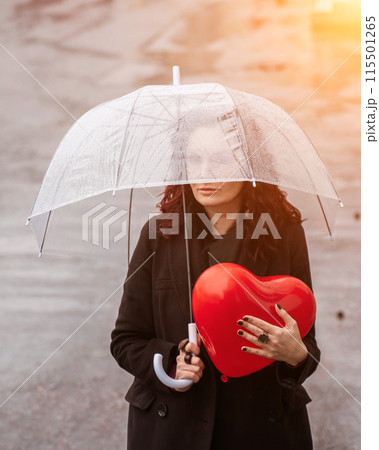 Sad middle-aged woman in a black coat and boots under a transparent umbrella. He holds a balloon in the form of a heart in his hands. Rain in the city, outdoor scene, water drops on the umbrella. The 115501265