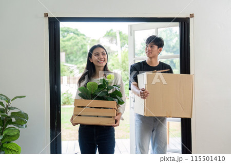Happy Couple Moving into New House, Carrying Boxes and Plants, Fresh Start in Modern Home, Relocation and New Beginnings, Young Adults Unpacking and Settling in Bright and Airy Living Space 115501410