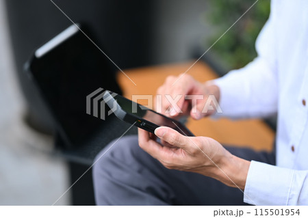 Young businessman typing text message on mobile phone while sitting outside office building Young businessman typing text message on mobile phone while sitting outside office building 115501954