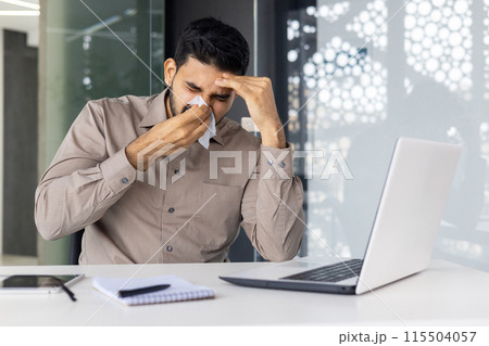 A sick young Indian man sits in the office at the desk and wipes his nose with a napkin from a runny nose, he does not feel well. A sick young Indian man sits in the office at the desk and wipes his nose with a napkin from a runny nose, he does not feel well. 115504057