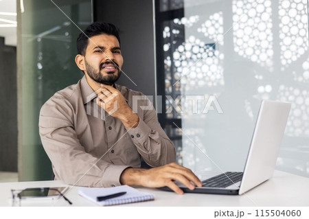 A young Indian man fell ill at work, sits at his desk, works on a laptop, holds his hand to his neck, feels a strong pain in his throat. A young Indian man fell ill at work, sits at his desk, works on a laptop, holds his hand to his neck, feels a strong pain in his throat. 115504060