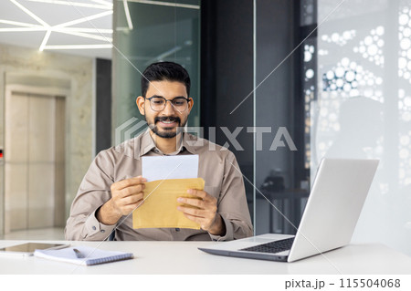 Smiling indian young man in glasses and shirt sitting in office at desk with laptop and reading received letter and document in open envelope. 115504068