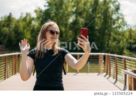 Active woman speaks via video link through the phone, takes a selfie near lake on nature background. Concept of an active lifestyle. Summer park 115505224