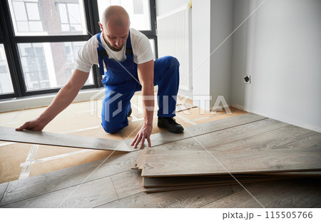 Male construction worker laying laminate wooden planks on floor underlayment. Man in work overalls installing laminate wood flooring in new flat. Home renovation concept. Male construction worker laying laminate wooden planks on floor underlayment. Man in work overalls installing laminate wood flooring in new flat. Home renovation concept. 115505766