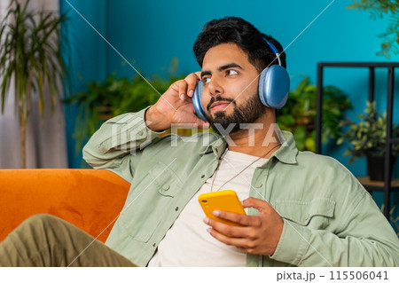 Happy relaxed man in wireless headphones dancing listening favorite disco pop music on home couch Happy relaxed man in wireless headphones dancing listening favorite disco pop music on home couch 115506041