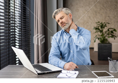 Mature man in blue shirt suffering from neck pain while sitting at desk and working on laptop in modern office. Mature man in blue shirt suffering from neck pain while sitting at desk and working on laptop in modern office. 115510943
