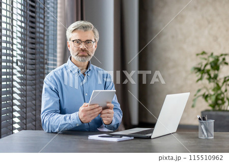 Mature businessman in modern office setting, using a digital tablet. Professional workspace with laptop, blinds, and indoor plants. 115510962