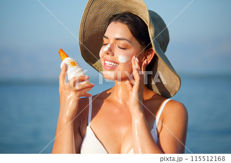 Beautiful Young woman with sun cream on face. Girl holding sunscreen bottle on the beach. Female in hat applying moisturizing lotion on skin.Skin care. Sun protection. Suntan 115512168