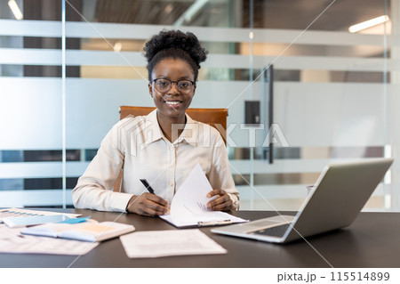 Confident businesswoman in modern office sitting at her desk with a laptop and documents, smiling while working. 115514899