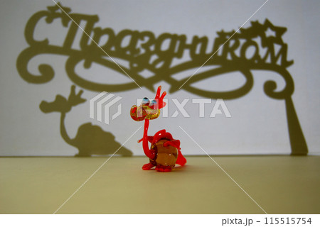 A toy dragon, its shadow and a shadow with an inscription. A little dragon and a congratulatory inscription Happy Holidays in Russian. 115515754