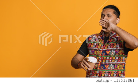 Sleepy man yawning and rubbing eyes. sipping coffee to wake up and gain energy. Exhausted person feeling fatigued after sleepless night, drinking caffeinated beverage, studio backdrop, camera A 115515861