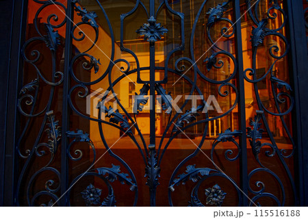 Beautiful, forged gate with many curls and leaves.Through the gate you can see the courtyard, illuminated by sunlight. Beautiful, forged gate with many curls and leaves.Through the gate you can see the courtyard, illuminated by sunlight. 115516188