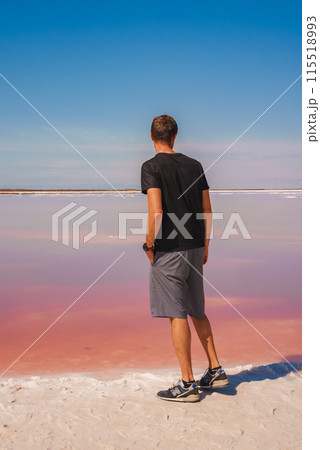 Person in casual attire stands at edge of pink lake under clear blue sky at Alviso Pink Lake Park, CA. Unique coloration creates serene, surreal scene. 115518993