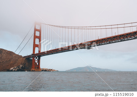 Iconic Golden Gate Bridge in SF, CA. View from water level shows the orange bridge against overcast sky. Tall towers, Marin Headlands visible. Serene scene. 115519010