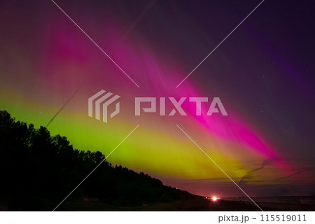 A stunning display of the aurora borealis illuminates the sky with green and pink hues over a coastal area near Riga and Jurmala, Latvia. A forest silhouette contrasts the vibrant colors. A stunning display of the aurora borealis illuminates the sky with green and pink hues over a coastal area near Riga and Jurmala, Latvia. A forest silhouette contrasts the vibrant colors. 115519011