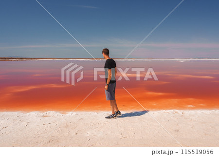 Person in black t shirt and grey shorts standing near pink lake admiring the vivid colors. Location Alviso Pink Lake Park, California. Sky is clear blue. Person in black t shirt and grey shorts standing near pink lake admiring the vivid colors. Location Alviso Pink Lake Park, California. Sky is clear blue. 115519056