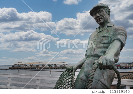 Bronze statue of a fisherman in action holding a net near ocean pier with cloudy sky. Depicts maritime culture in coastal town or fishing destination. 115519140