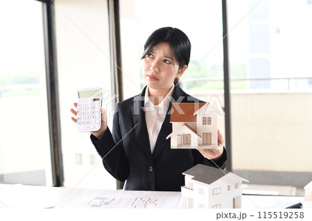 住宅模型と電卓を持つスーツ姿の悩む女性 住宅模型と電卓を持つスーツ姿の悩む女性 115519258