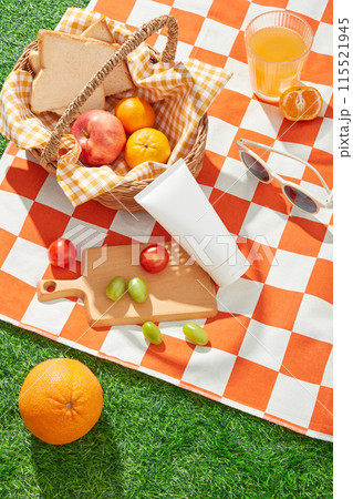 Scene of summertime on green meadow, an orange carpet displays a rattan basket of picnic food, orange, glasses and wooden tray. Mock up for advertising with blank label tube Scene of summertime on green meadow, an orange carpet displays a rattan basket of picnic food, orange, glasses and wooden tray. Mock up for advertising with blank label tube 115521945
