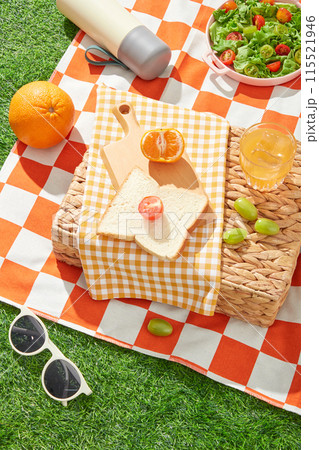 Creative template of picnic theme on grass, from top view. A yellow towel spread on a rattan basket with a wooden tray of sandwich and fruit above, below is an orange carpet contains picnic items Creative template of picnic theme on grass, from top view. A yellow towel spread on a rattan basket with a wooden tray of sandwich and fruit above, below is an orange carpet contains picnic items 115521946