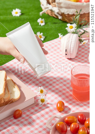 Cosmetics advertising photo with a picnic concept from a high angle, an empty label tube is held by a beautiful hand, below is a pink plaid rug containing some picnic items 115521947