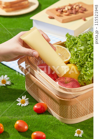 The unlabeled cosmetic tube in yellow color picked out of a wicker basket, which placed on green grass and containing some fresh vegetable and fruit for picnic meal. Photo of high angle, copy space The unlabeled cosmetic tube in yellow color picked out of a wicker basket, which placed on green grass and containing some fresh vegetable and fruit for picnic meal. Photo of high angle, copy space 115521948