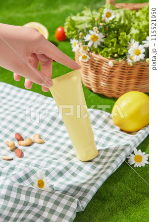 A yellow blank label cosmetic tube has just been placed on a blue carpet with a checkered pattern, behind is a green lawn, a rattan basket filled with feverfew flowers and a yellow lemon 115521950