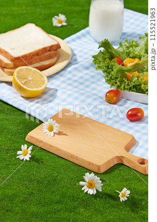 Summer picnic concept photo with high angle. On the green grass lay a checkered blue carpet, sandwiches, salad, a bottle of milk and an empty tray placed on top. Product presentation space Summer picnic concept photo with high angle. On the green grass lay a checkered blue carpet, sandwiches, salad, a bottle of milk and an empty tray placed on top. Product presentation space 115521963