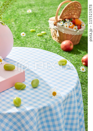 A table with a blue tablecloth, has a few grapes scattered on top. In the distance is a rattan basket containing fruit placed on the grass. Photo of high angle with empty space for displaying 115521978