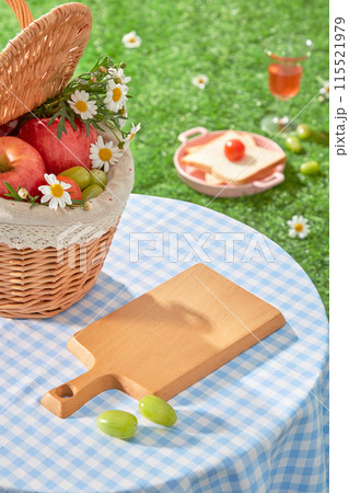 Outdoor picnic photo from above shot, a blank wooden tray placed on a checkered blue table amidst the grass, next to a rattan basket full of fruit. Vacant space for adding design elements 115521979