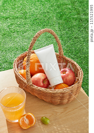On the table displays a rattan picnic basket of fruit, with a white blank label tube of cosmetic above, next to a glass of orange juice. High angle shot photo on grass surface, space for text On the table displays a rattan picnic basket of fruit, with a white blank label tube of cosmetic above, next to a glass of orange juice. High angle shot photo on grass surface, space for text 115521995