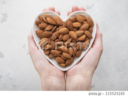 Female hands holding heart shaped plate with peeled almond nuts on light background. Female hands holding heart shaped plate with peeled almond nuts on light background. 115525285