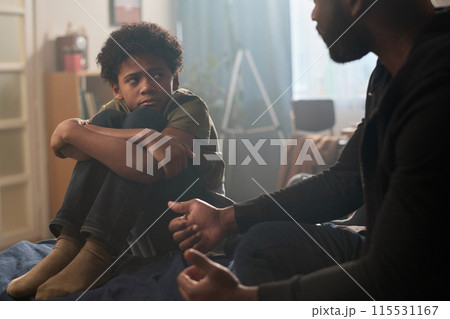 Full length portrait of troubled teenage boy talking to father sitting on bed and hugging knees copy space 115531167