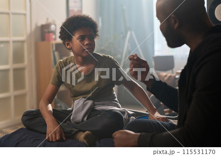 Portrait of young Black boy talking to father sitting on bed together in affectionate setting Portrait of young Black boy talking to father sitting on bed together in affectionate setting 115531170