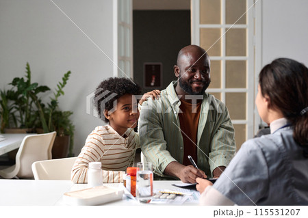Portrait of smiling African American father and son signing medical form with female doctor giving instructions during home visit copy space 115531207