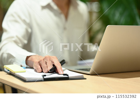Cropped shot of businessman checking financial data in report and using laptop on wooden desk Cropped shot of businessman checking financial data in report and using laptop on wooden desk 115532392