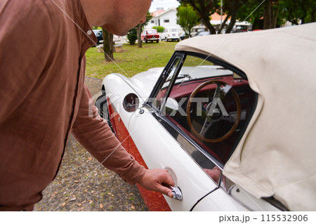 A stylish man in a shirt holds on to the handle of a retro car. 115532906