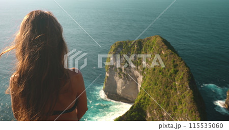 A woman stands confidently on top of a cliff overlooking the ocean on Nusa Penida Island in Indonesia. The powerful waves crash against the rocks below as she gazes out at the vast blue sea. 115535060