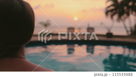 A woman in a swimsuit is standing by the edge of a swimming pool in Thailand. The water is still, reflecting the surrounding palms and sky. 115535088