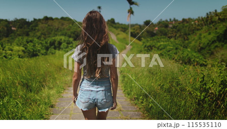 Woman walking Campuhan Ridge Walk Trail, Indonesia, Bali island, Ubud. Back view long hair tourist girl enjoy green hill nature landscape. 115535110