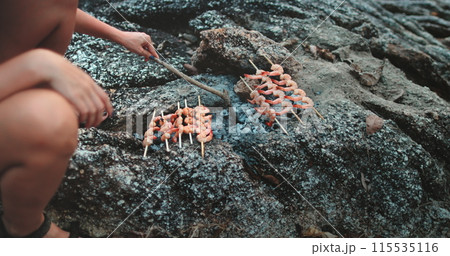 A person is sitting on a large rock while holding and eating food skewered on sticks, likely a seafood barbecue dish. The individual appears relaxed and enjoying the outdoor dining experience. A person is sitting on a large rock while holding and eating food skewered on sticks, likely a seafood barbecue dish. The individual appears relaxed and enjoying the outdoor dining experience. 115535116