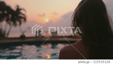 Woman relaxes near swimming pool at sunset. Long hair brunette girl back view enjoys colorful sky reflected in water, palm trees in background. Tropical resort, travel, exotic vacation in Thailand 115535120