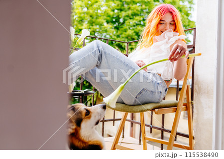 Playful woman with pink hair is sitting on a chair with a flower in her hand. A dog is standing behind her, looking at her. The scene is peaceful and relaxing, female enjoying time outdoors Playful woman with pink hair is sitting on a chair with a flower in her hand. A dog is standing behind her, looking at her. The scene is peaceful and relaxing, female enjoying time outdoors 115538490